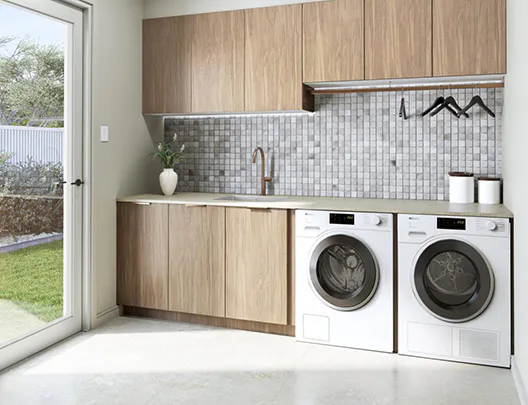 Contemporary laundry renovation featuring warm timber cabinetry, mosaic tile splashback, front-loading washer and dryer, and a large glass door opening to a sunny backyard.