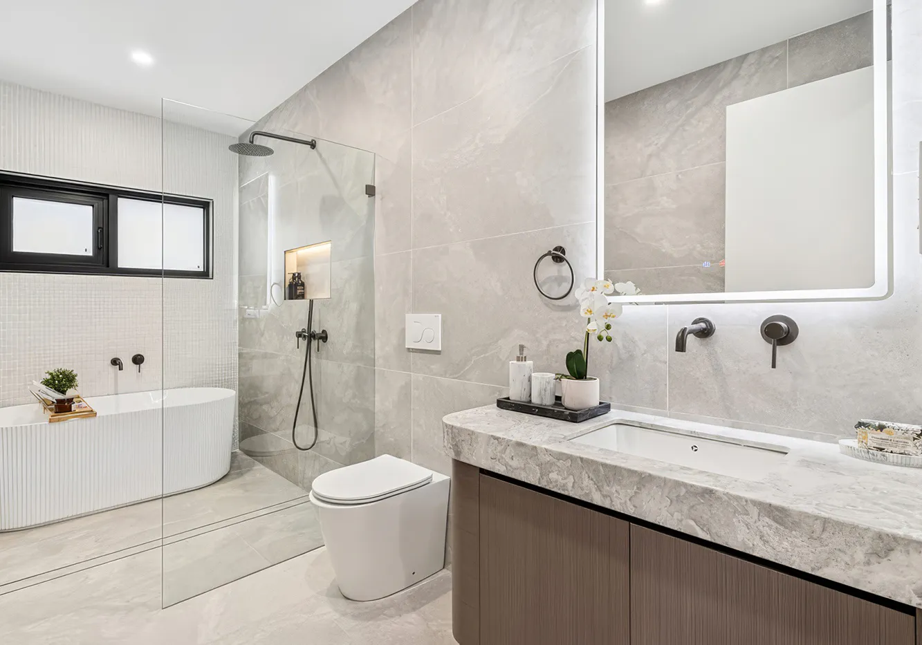 Modern bathroom renovation featuring a freestanding bathtub, walk-in shower with rainfall showerhead, wall-mounted vanity with stone benchtop, and backlit mirror — neutral tones and contemporary fittings creating a sleek, spa-like finish.